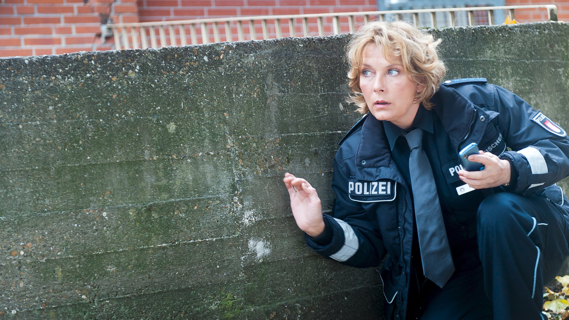 "Notruf Hafenkante - Figaros Rache": Claudia (Janette Rauch) kniet geduckt neben einer Mauer und scheint etwas zu beobachten.