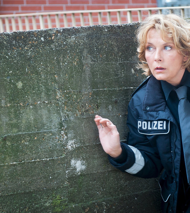"Notruf Hafenkante - Figaros Rache": Claudia (Janette Rauch) kniet geduckt neben einer Mauer und scheint etwas zu beobachten.