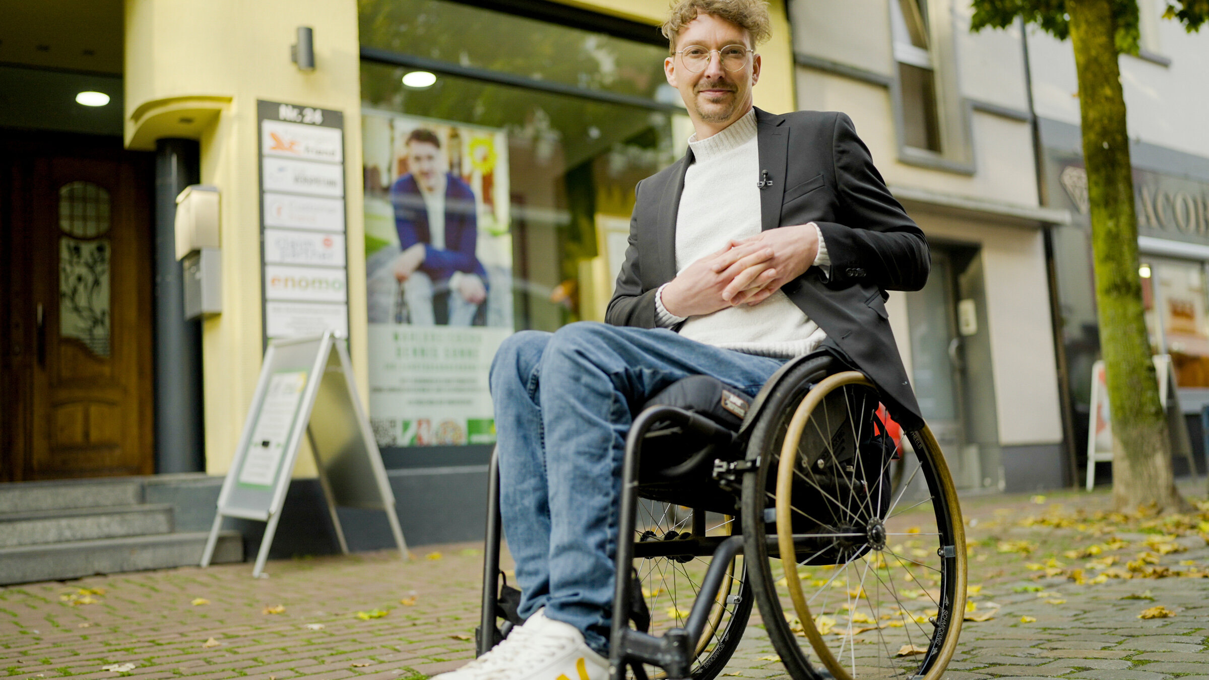 "Einfach Mensch - Vom Sehen und Gesehen werden": Dennis Sonne sitzt in seinem Rollstuhl in der Stadt vor seinem Wahlkreisbüro. Er schaut lächelnd in die Kamera.