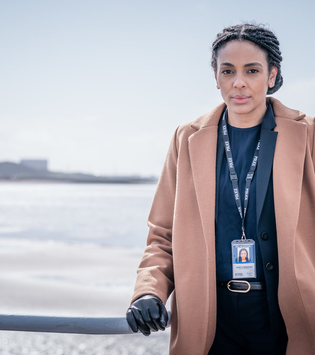 "The Bay - Neustart": DS Jenn Townsend (Marsha Thomason) lehnt sich an ein Geländer und schaut dabei konzentriert in die Kamera. Im Hintergrund ist das raue Meer zu erkennen, während am Horizont die Umrisse von einigen Gebäuden zu sehen sind.