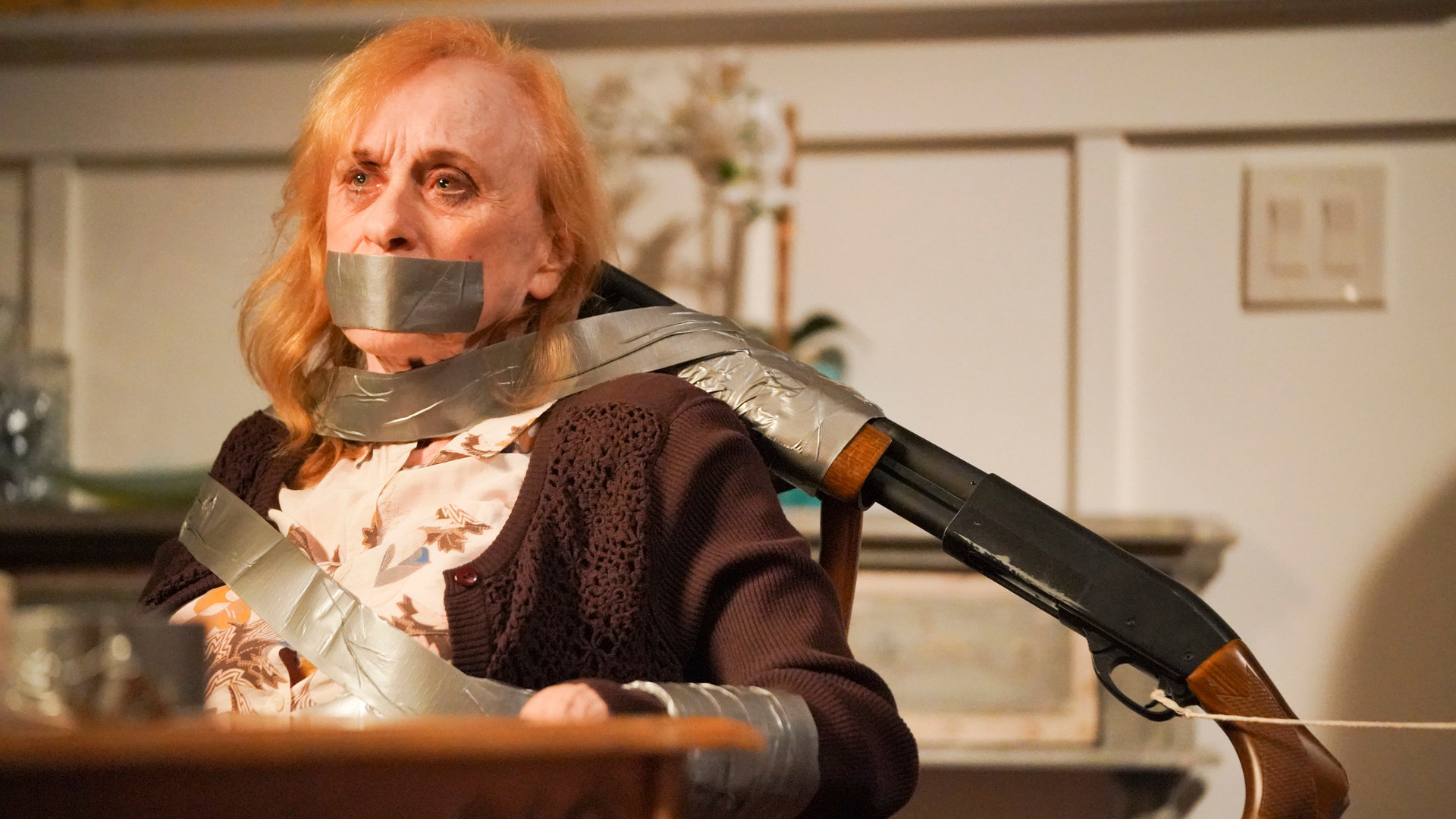 "The Rookie - Menschenjagd": Sallie (Jeannie Epper) ist mit Paketband an einen Stuhl gefesselt. Sie hat einen ängstlichen Gesichtsausdruck. Ein Gewehr ist mit Tape an der Stuhllehne befestigt und auf Sallies Kopf gerichtet.