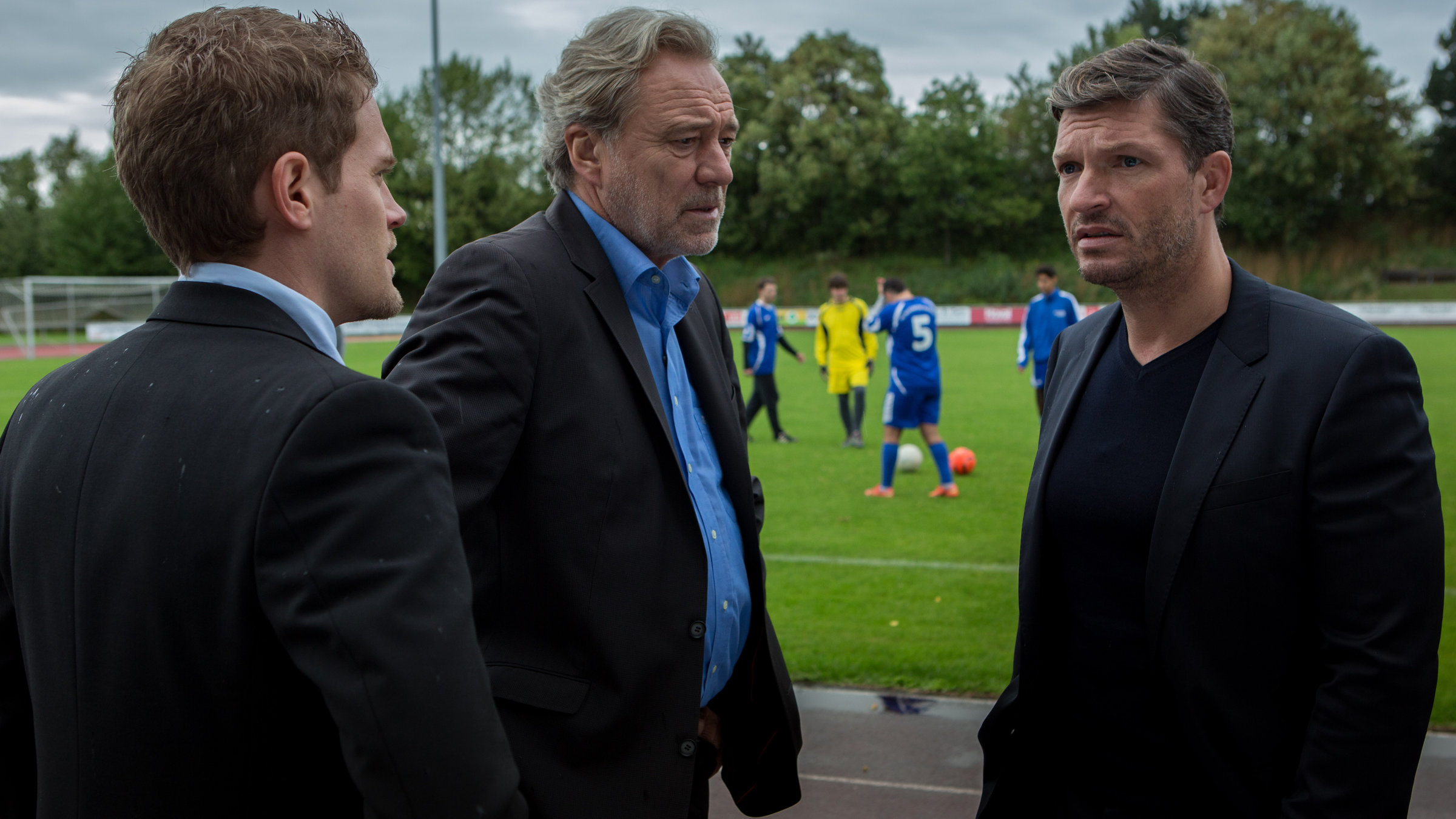 "SOKO München - Scheiß Schiri": Auf dem Fußballplatz befragen Dominik Morgenstern (Joscha Kiefer) und Arthur Bauer (Gerd Silberbauer) Thomas Grimm (Hardy Krüger jr.), sie stehen am Rand des Fußballfeldes. Auf dem Platz sind Spieler beim Training zu sehen.