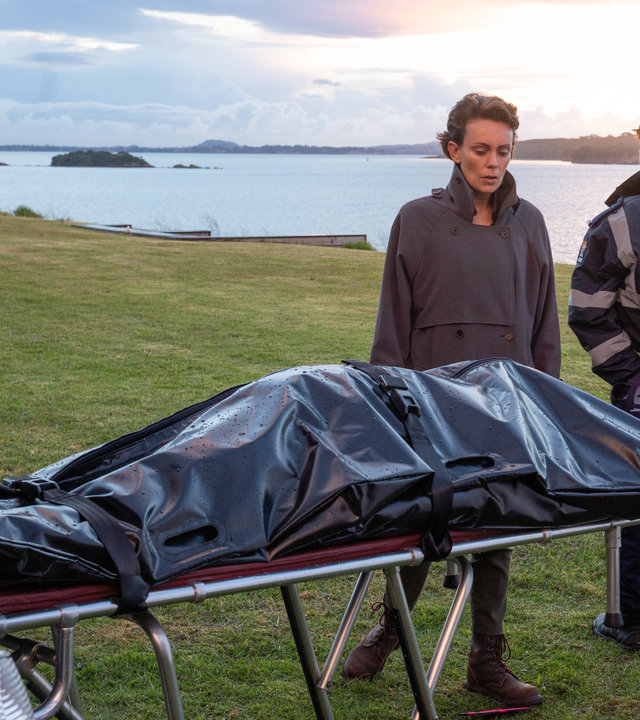 "Auckland Detectives - Tödliche Bucht: Gefährliche Nähe": Detective Senior Sergeant Jessica Savage (Kate Elliott) steht auf einer Wiese am Meer hinter einem Leichenwagen vor einer Bahre und schaut auf einen schwarzen Leichensack. Neben ihr, den Rücken zu ihr gekehrt, steht ein Streifenpolizist (Darsteller unbekannt), der seine Dienstmütze hinter seinem Rücken in der Hand hält.