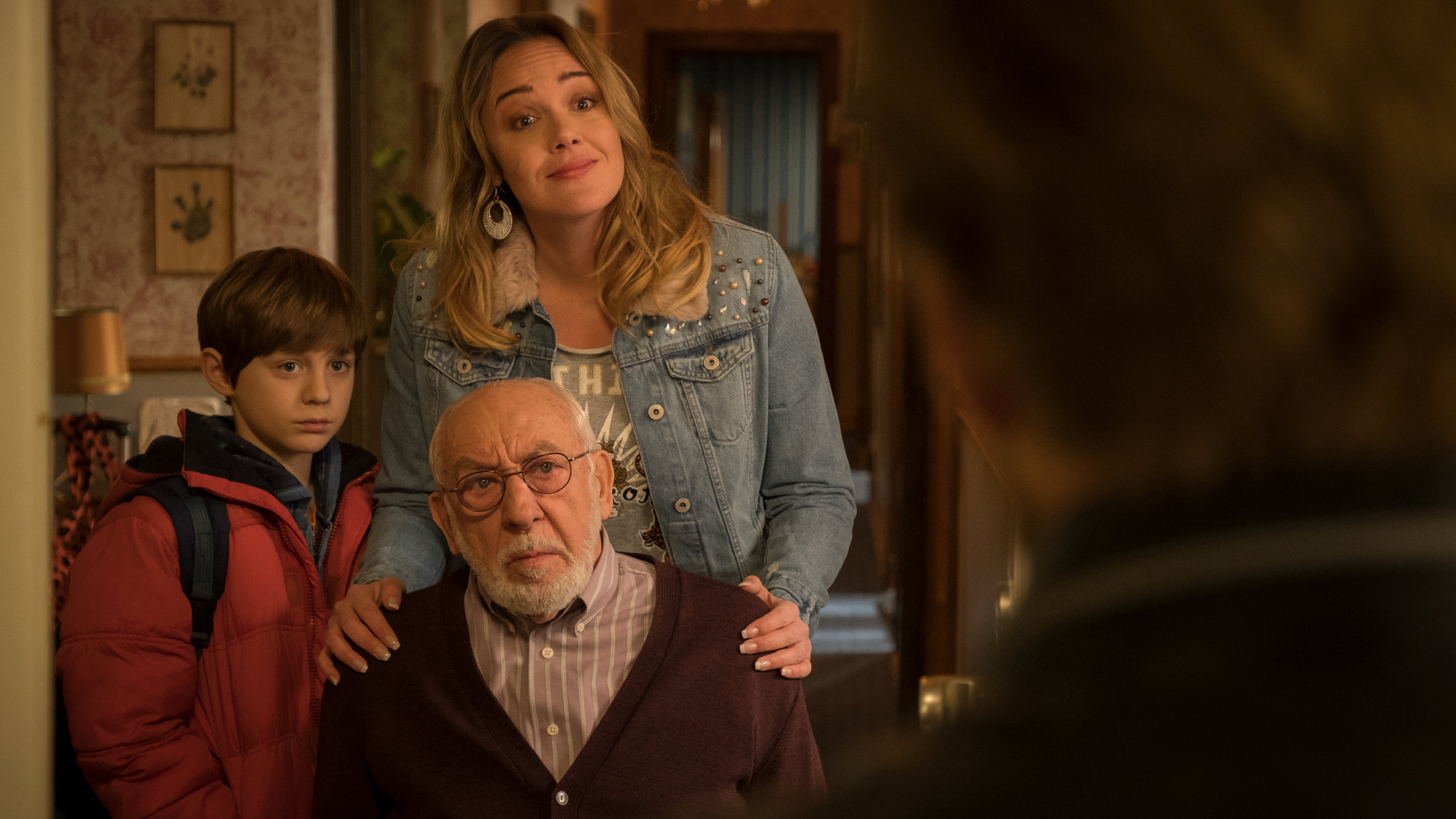 "Mein Freund, das Ekel": Hintz (Dieter Hallervorden) sitzt im Rollstuhl. Hinter ihm steht, ihre Hände auf seinen Schultern, Trixie (Alwara Höfels) und ihr Sohn Sean (Lior Kudrjawizki). Alle schauen zu einer Person, die mit dem Rücken zur Kamera steht.