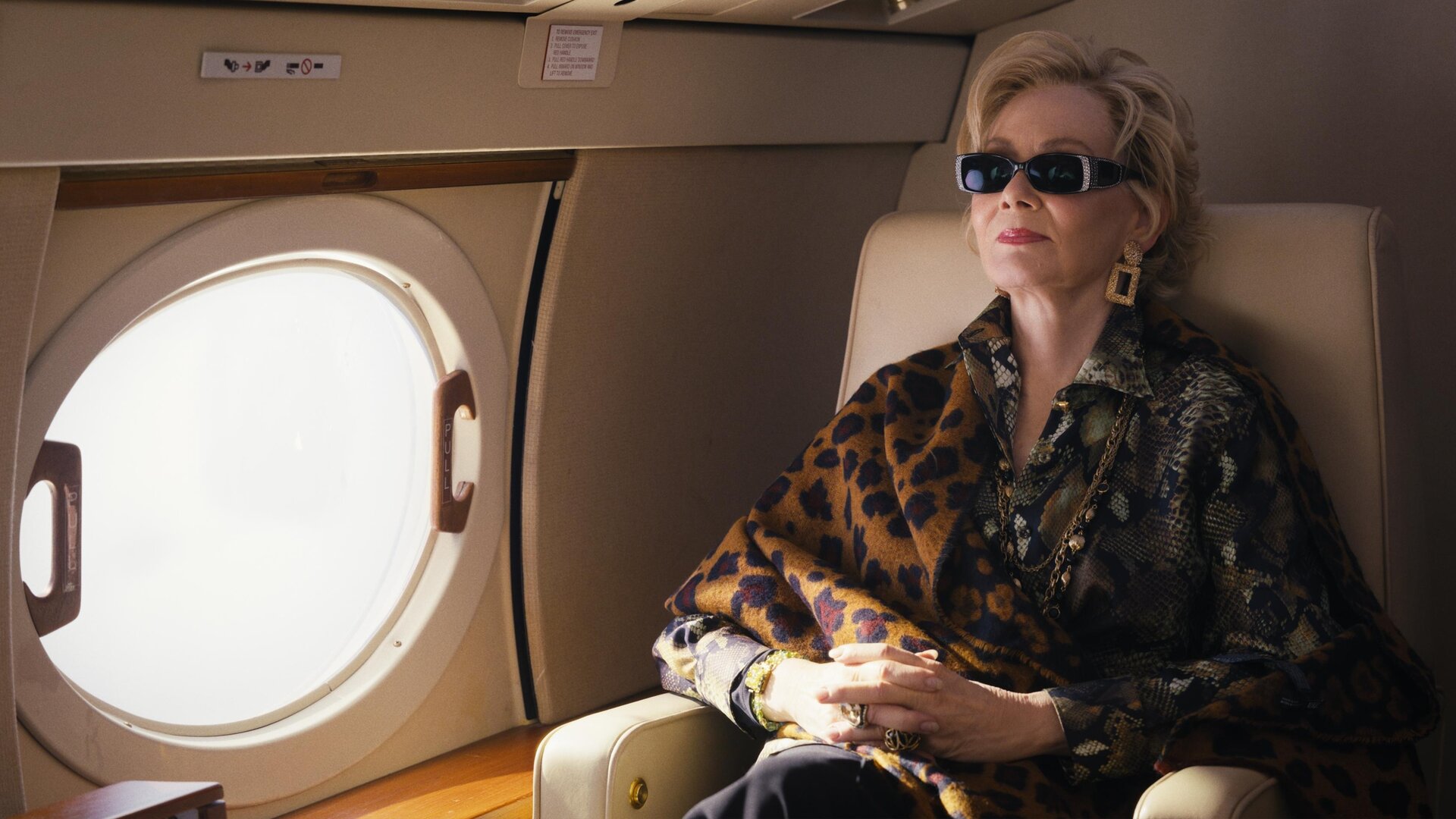 "Hacks - Willkommen im Club": Deborah sitzt mit Sonnenbrille, goldenen Ohrringen und Ketten und mit gefalteten Händen in einem Sessel ihres Privatjets. Neben ihr ist ein Fenster des Flugzeugs zu sehen.