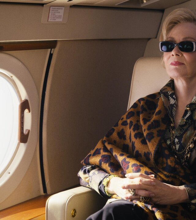 "Hacks - Willkommen im Club": Deborah sitzt mit Sonnenbrille, goldenen Ohrringen und Ketten und mit gefalteten Händen in einem Sessel ihres Privatjets. Neben ihr ist ein Fenster des Flugzeugs zu sehen.