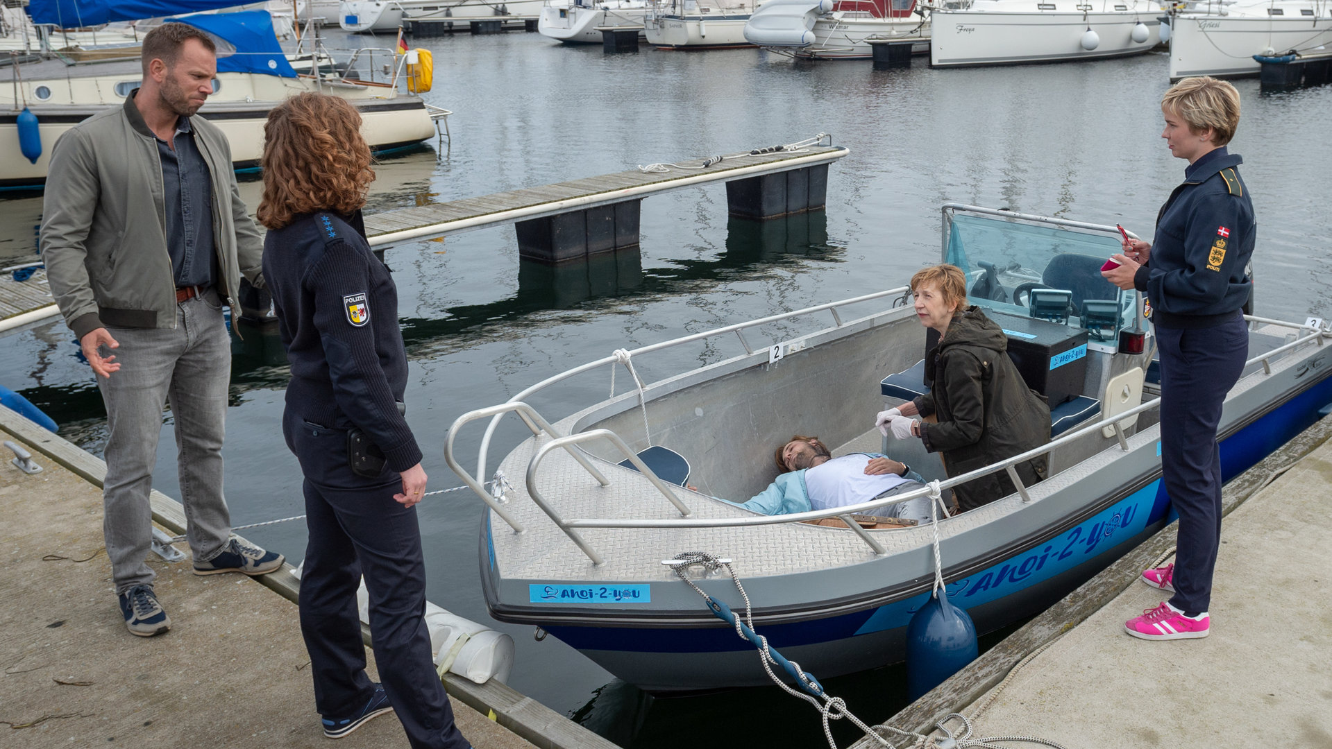 "SOKO Wismar – Tod ahoi!": Im Yachthafen liegt das Opfer in einem Boot, Helene Sturbeck (Katharina Blaschke) kniet neben ihm. Auf einer Seite des Stegs stehen Lars Pöhlmann (Dominic Boeer) und Paula Moorkamp (Stella Hinrichs) - auf der anderen Seite stehen Stine Bergendal (Sidsel Hindhede), Roswitha Prinzler (Silke Matthias) und weitere Kolleginnen und Kollegen (Komparsen).