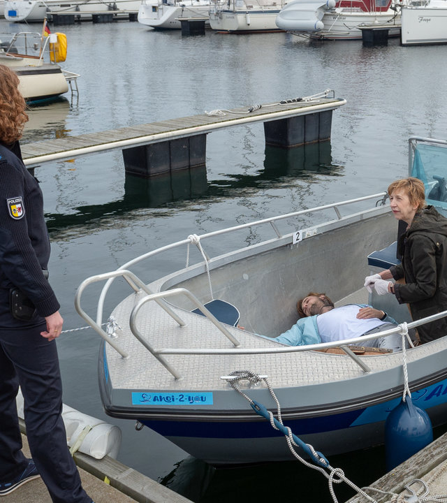 "SOKO Wismar – Tod ahoi!": Im Yachthafen liegt das Opfer in einem Boot, Helene Sturbeck (Katharina Blaschke) kniet neben ihm. Auf einer Seite des Stegs stehen Lars Pöhlmann (Dominic Boeer) und Paula Moorkamp (Stella Hinrichs) - auf der anderen Seite stehen Stine Bergendal (Sidsel Hindhede), Roswitha Prinzler (Silke Matthias) und weitere Kolleginnen und Kollegen (Komparsen).