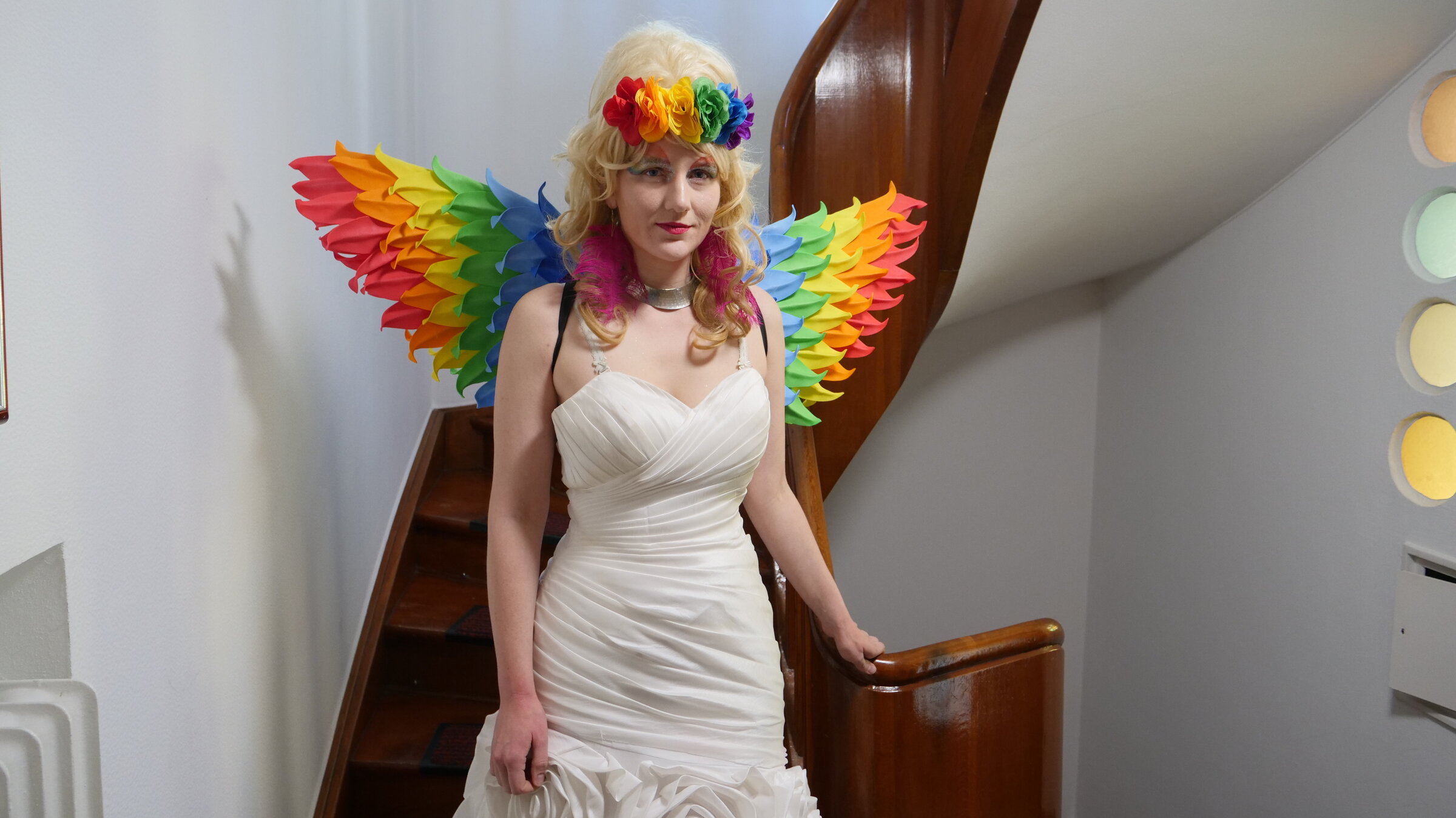 "37°Leben: Mut unterm Kreuz: Leben im Kloster": Schwester Micha steht vor einer Treppe mit einem Kirchenfenster. Sie trägt ein weißes Kleid, eine blonde Perücke und Flügel in Regenbogenfarben. Sie ist grell geschminkt.