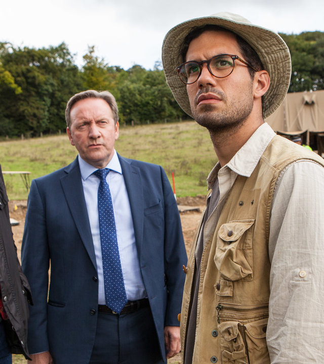 "Inspector Barnaby - Heilige und Eilige": An einer archäologischen Grabungsstätte stehen DS Nelson (Gwilym Lee) und DCI Barnaby (Neil Dudgeon) nebeneinander. Sie schauen zu dem Grabungsassistenten Dexter Ingram (Stefano Braschi), der vor ihnen steht und seinen Blick abgewandt hat.