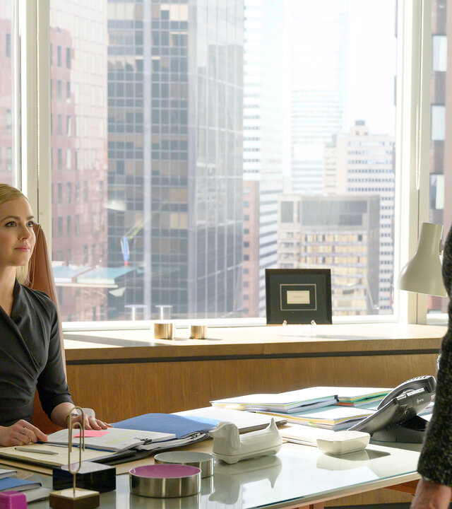 "Suits - Zwangsverwaltung": Katrina (Amanda Schull) sitzt am Schreibtisch, auf dem etliche Akten und Dokumente ausgebreitet liegen. Vor ihr steht Faye (Denise Crosby), die ihre Hände auf zwei Stühle neben ihr gelegt hat.