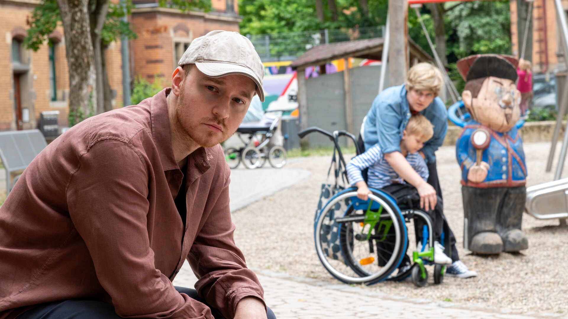 "SOKO Stuttgart - Gefährliches Gespann": Jakub Piontek (Wiktor Grduszak, l.) sitzt auf der Bank eines öffentlichen Spielplatzes. Hinter ihm sind eine Frau (Komparsin, r.) und ein kleiner Junge (Komparse) im Rollstuhl zu sehen. Die Frau hilft dem Kind beim Einsteigen in den Rollstuhl. Sie schaut in Pionteks Richtung, doch er dreht den Kopf weg, als wolle er nicht erkannt werden.