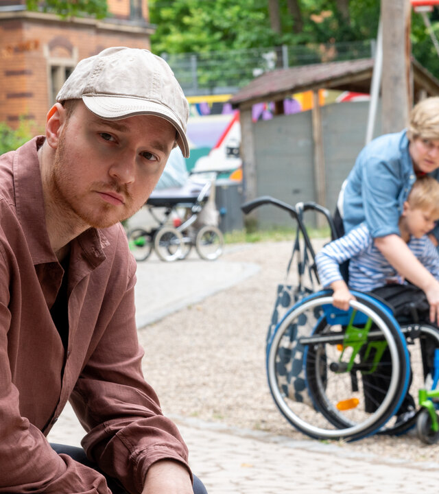 "SOKO Stuttgart - Gefährliches Gespann": Jakub Piontek (Wiktor Grduszak, l.) sitzt auf der Bank eines öffentlichen Spielplatzes. Hinter ihm sind eine Frau (Komparsin, r.) und ein kleiner Junge (Komparse) im Rollstuhl zu sehen. Die Frau hilft dem Kind beim Einsteigen in den Rollstuhl. Sie schaut in Pionteks Richtung, doch er dreht den Kopf weg, als wolle er nicht erkannt werden.