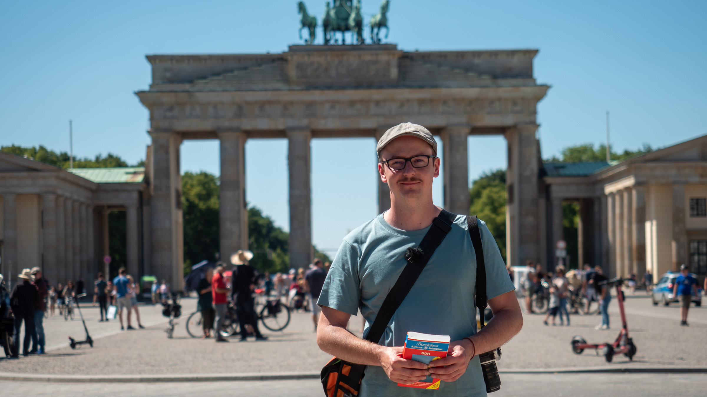 "Lost in DDR - Stefan Danzigers Trip nach 1989": Auf dem Bild steht Stefan Danziger vor dem Brandenburger Tor in Berlin. Er nimmt den Mittelpunkt des Bildes ein. Danziger hat eine orangene Tasche umhängen und hält in seinen Händen einen alten DDR-Reiseführer. Er trägt ein blaues T-Shirt, eine Brille und eine Mütze. Hinter dem Brandenburger Tor sieht man strahlend blauen Himmel.