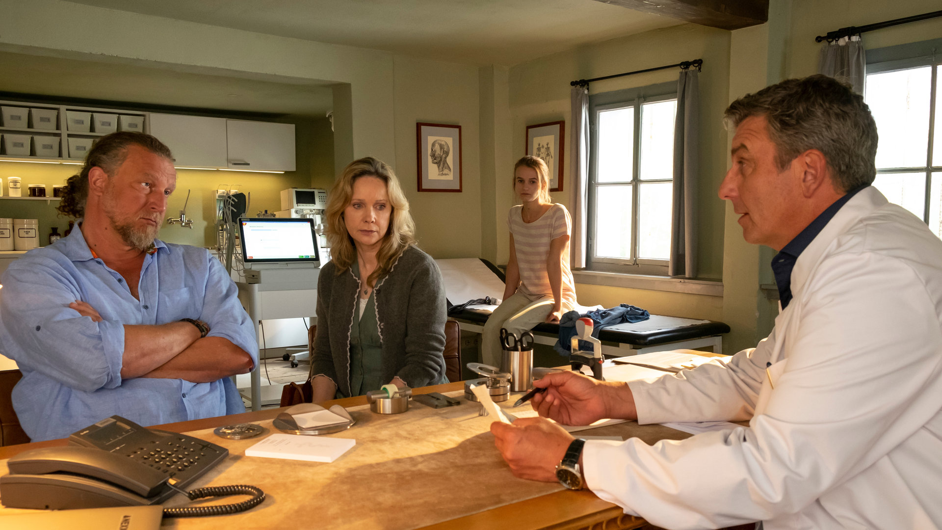 "Der Bergdoktor: Endlich Leben": Dr. Martin Gruber (Hans Sigl) sitzt mit ernster Miene vor Gero (Matthias Komm) und Maren Engl (Ann-Kathrin Kramer) an seinem Schreibtisch. Chiara Engl (Leonie Wesselow) sitzt im Hintergrund auf der Krankenliege.