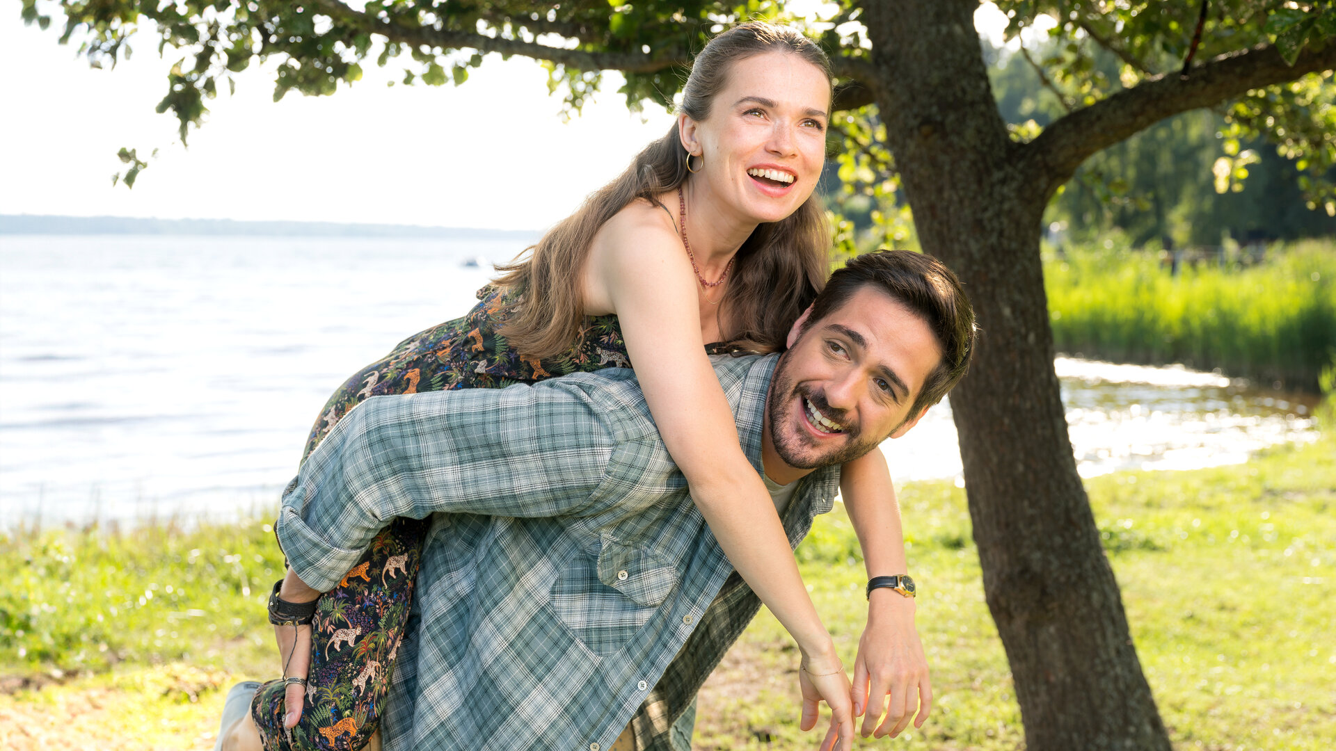 "Inga Lindström: Herz über Kopf": Nora (Zoe Moore) und Samu (Félix Herzog) befinden sich im Freien unter einem Baum am Ufer eines Gewässers. Samu hat Nora Huckepack genommen, beide schauen lachend in die sonnige Landschaft.