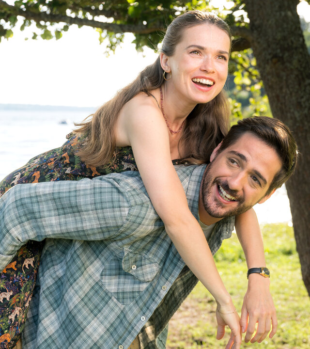 "Inga Lindström: Herz über Kopf": Nora (Zoe Moore) und Samu (Félix Herzog) befinden sich im Freien unter einem Baum am Ufer eines Gewässers. Samu hat Nora Huckepack genommen, beide schauen lachend in die sonnige Landschaft.