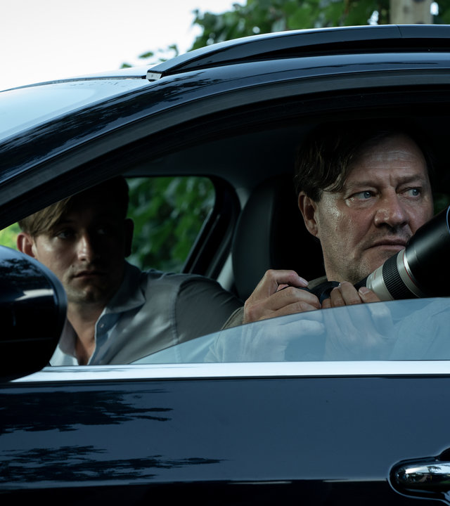 "Im Netz der Camorra": Silvio Bernatti (Lukas Watzl) und Adrin Erlacher (Harald Windisch) sitzen in einem Auto und schauen aus dem Fenster der Fahrerseite in die gleiche Richtung. Erlacher hat eine Kamera mit Zoomobjektiv in der Hand.
