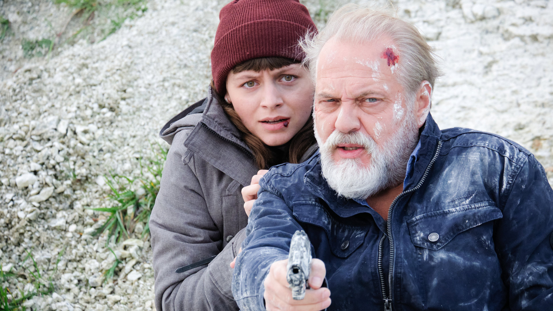"Der Alte - Tod am Kliff": Paula Voss (Antonia Breidenbach) versteckt sich hinter der Schulter ihres Vaters Richard Voss (Jan-Gregor Kremp), der mit einer Schürfwunde am Kopf und mit gezogener Pistole in der Hand an einen Abhang mit Schutt gelehnt sitzt.