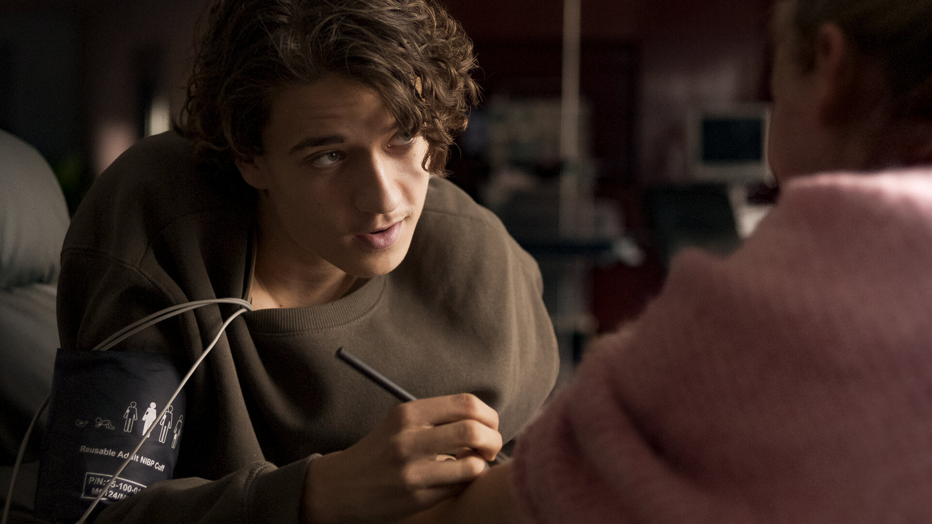 "Doktor Ballouz - Erste Liebe": Ronni (Marven Gabriel Suarez-Brinkert) sitzt Juna (Lina Hüesker) gegenüber, die nur im Anschnitt von hinten zu sehen ist. Er schreibt seine Nummer auf ihren Arm.