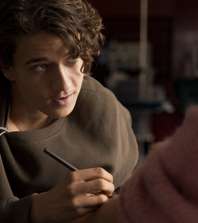 "Doktor Ballouz - Erste Liebe": Ronni (Marven Gabriel Suarez-Brinkert) sitzt Juna (Lina Hüesker) gegenüber, die nur im Anschnitt von hinten zu sehen ist. Er schreibt seine Nummer auf ihren Arm.