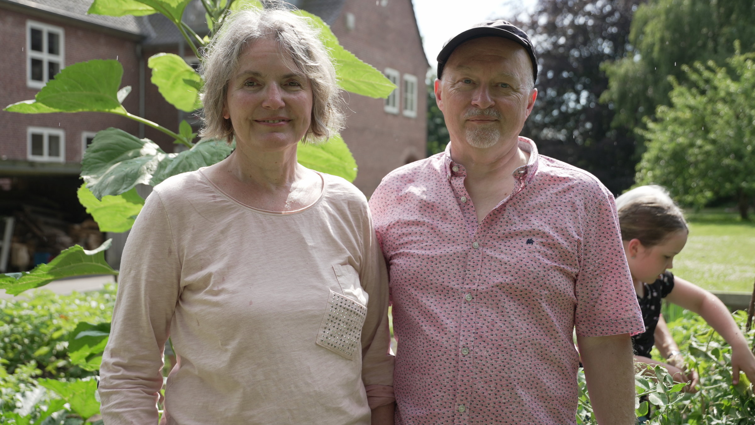 "37° Seniorenheim oder Wohnprojekt? - Neustart mit 60+": Die Eheleute Martina und Thomas stehen im Garten vor dem Haus. Sie schauen beide lächelnd in die Kamera.