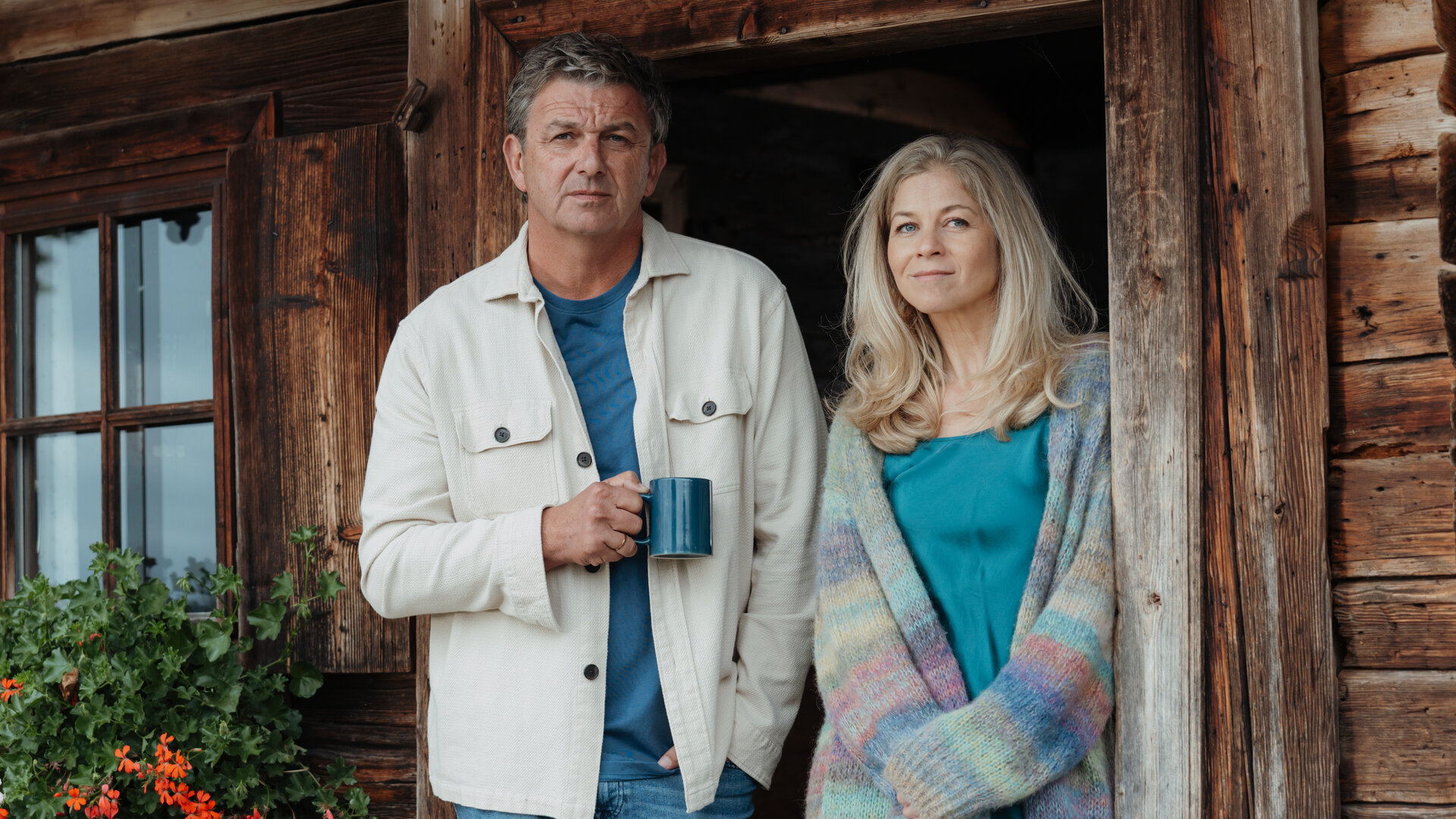 "Der Bergdoktor: Schmelzpunkte": Auf dem Gruberhof: In der geöffneten Tür steht Dr. Martin Gruber (Hans Sigl) und hält eine Kaffeetasse in der Hand. Neben ihm steht Karin Bachmeier (Hilde Dalik) mit offenem Haar und einer langen Strickjacke. Beide blicken lächelnd in die Ferne.