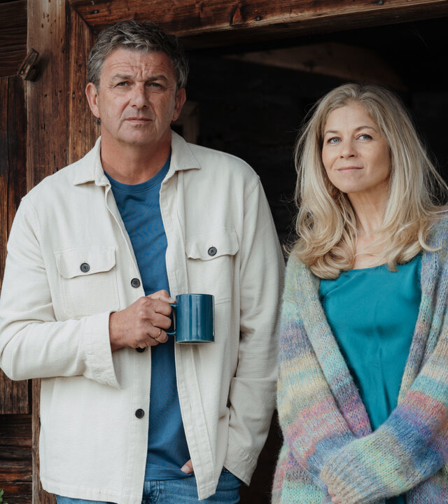 "Der Bergdoktor: Schmelzpunkte": Auf dem Gruberhof: In der geöffneten Tür steht Dr. Martin Gruber (Hans Sigl) und hält eine Kaffeetasse in der Hand. Neben ihm steht Karin Bachmeier (Hilde Dalik) mit offenem Haar und einer langen Strickjacke. Beide blicken lächelnd in die Ferne.