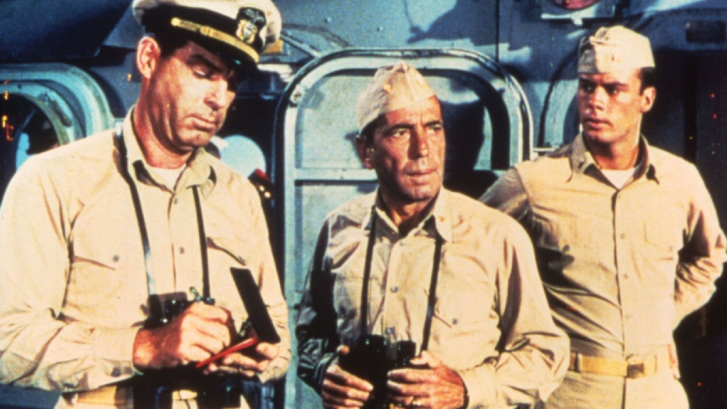 "Die Caine war ihr Schicksal": Vor den Aufbauten eines Schiffes steht ein Mann in Uniform  (Humphrey Bogart) mit einem umgehängten Fernglas. Rechts und links von ihm befinden sich zwei weitere Uniformierte, von denen der Mann links Eintragungen in eine Art Buch vornimmt, während der andere erstaunt blickt und die Hände in die Seiten stemmt.
