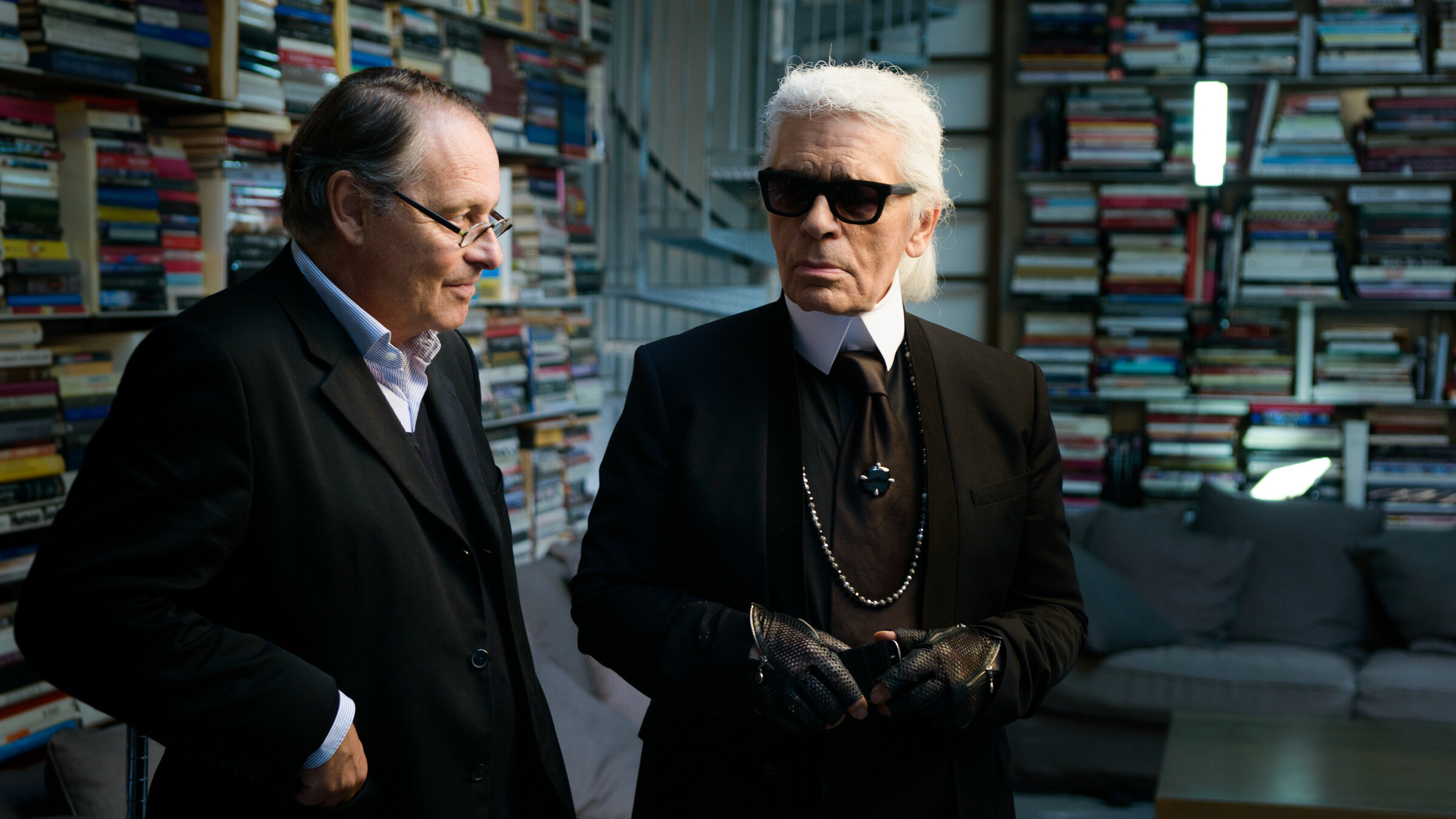"Karl - Der Mann hinter der Maske": Karl Lagerfeld und Gero von Boehm stehen in einem Raum voller Bücher. Lagerfeld trägt ein schwarzes Outfit mit Krawatte, Sonnenbrille, schwarzen Lederhandschuhen und einer langen Perlenkette. Sein weißes Haar ist streng nach hinten gekämmt. Gero von Boehm, neben ihm, trägt einen dunklen Anzug mit Brille und scheint ihm zuzuhören. Im Hintergrund sind hohe Regale voller bunter Bücher sowie eine Couch mit Kissen zu sehen.