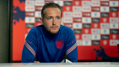 "Dear England (2)": Harry Kane (Will Antenbring) sitzt mit nachdenklichem Gesichtsausdruck an einem Tisch. Er trägt ein überwiegend blaues Trikot mit weiß-roten Streifen auf den Ärmeln und mit England-Emblem.