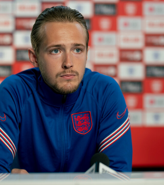 "Dear England (2)": Harry Kane (Will Antenbring) sitzt mit nachdenklichem Gesichtsausdruck an einem Tisch. Er trägt ein überwiegend blaues Trikot mit weiß-roten Streifen auf den Ärmeln und mit England-Emblem.