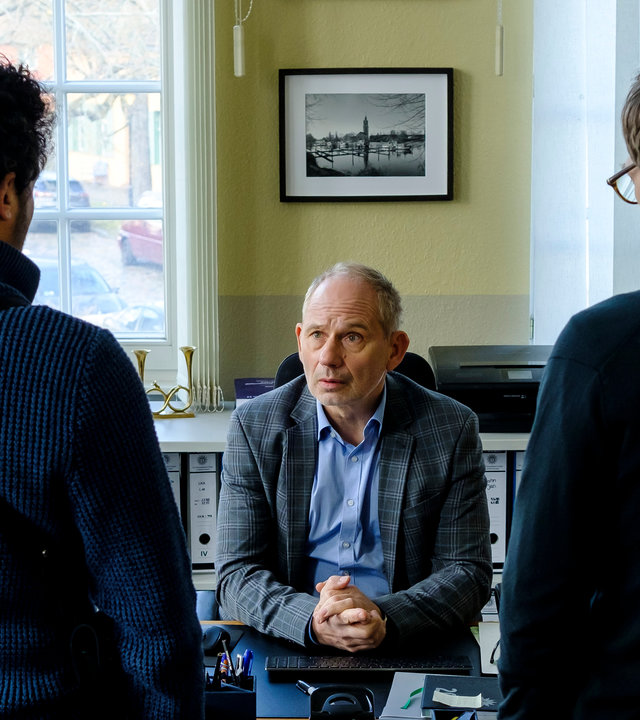 "SOKO Potsdam - Bittersüßer Tod": David Grünbaum (Omar El-Saeidi) und Christoph Westerman (Hendrik von Bültzingslöwen) stehen mit dem Rücken zur Kamera vor dem Schreibtisch ihres Chefs Bernhard Henschel (Michael Lott). Dieser schaut zu David.