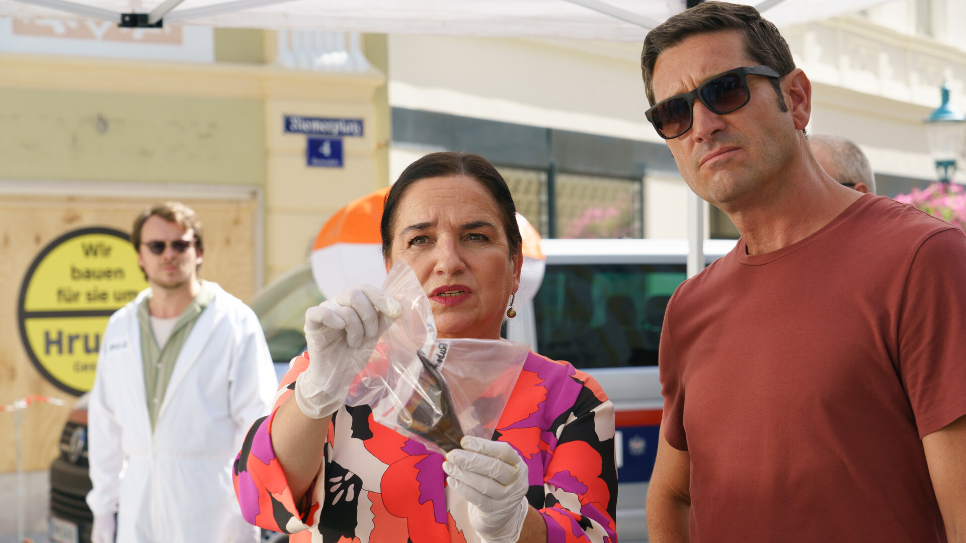 "SOKO Wien - Ausgelöscht": Dr. Franziska Beck (Maria Happel) steht im Freien und hält mit behandschuhten Händen einen dunklen Gegenstand in einer Plastiktüte in die Höhe. Klaus Lechner (Andreas Kiendl) steht neben ihr und trägt eine Sonnenbrille. Julius Rubatsch (Max Fischnaller) steht im weißen Anzug der Spurensicherung im Hintergrund und trägt ebenfalls eine Sonnenbrille.
