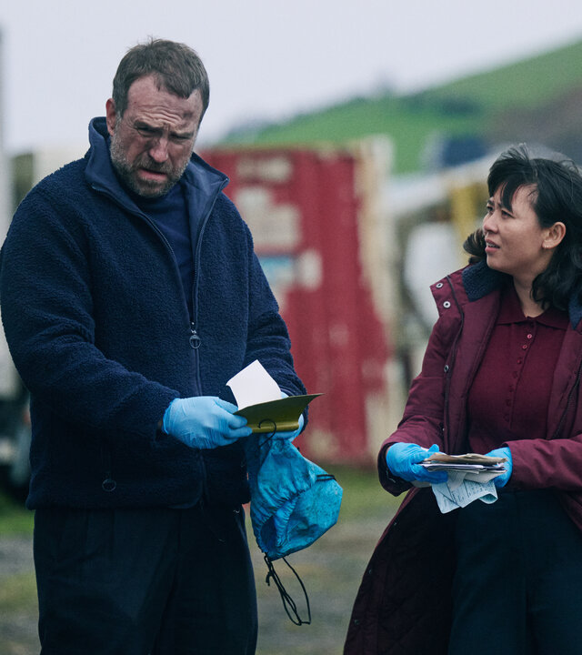 "Under Salt Marsh - Tod in den Salzwiesen - Im Auge des Sturms": Osian (Julian Lewis Jones) steht im Freien vor mehreren groÃen Fahrzeugen und hÃ¤lt ein kleines Buch in HÃ¤nden. Detective Jess Deng (Naomi Yang) steht neben ihm und blickt ihm an. Um ihn herum ist es dunkel.