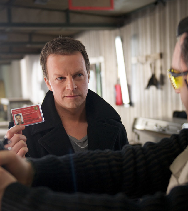 "SOKO Wismar - Schusslinie": In einem Raum des Schießvereins steht Vereinsmitglied Roth (Rainer Laupichler) mit Schutzbrille, gelben Ohrschützer und Schusswaffe in der Hand zur Schießwand gerichtet. Er redet dabei mit Sven Herzog (Michael Härle), der seinen Ausweis hochhält.