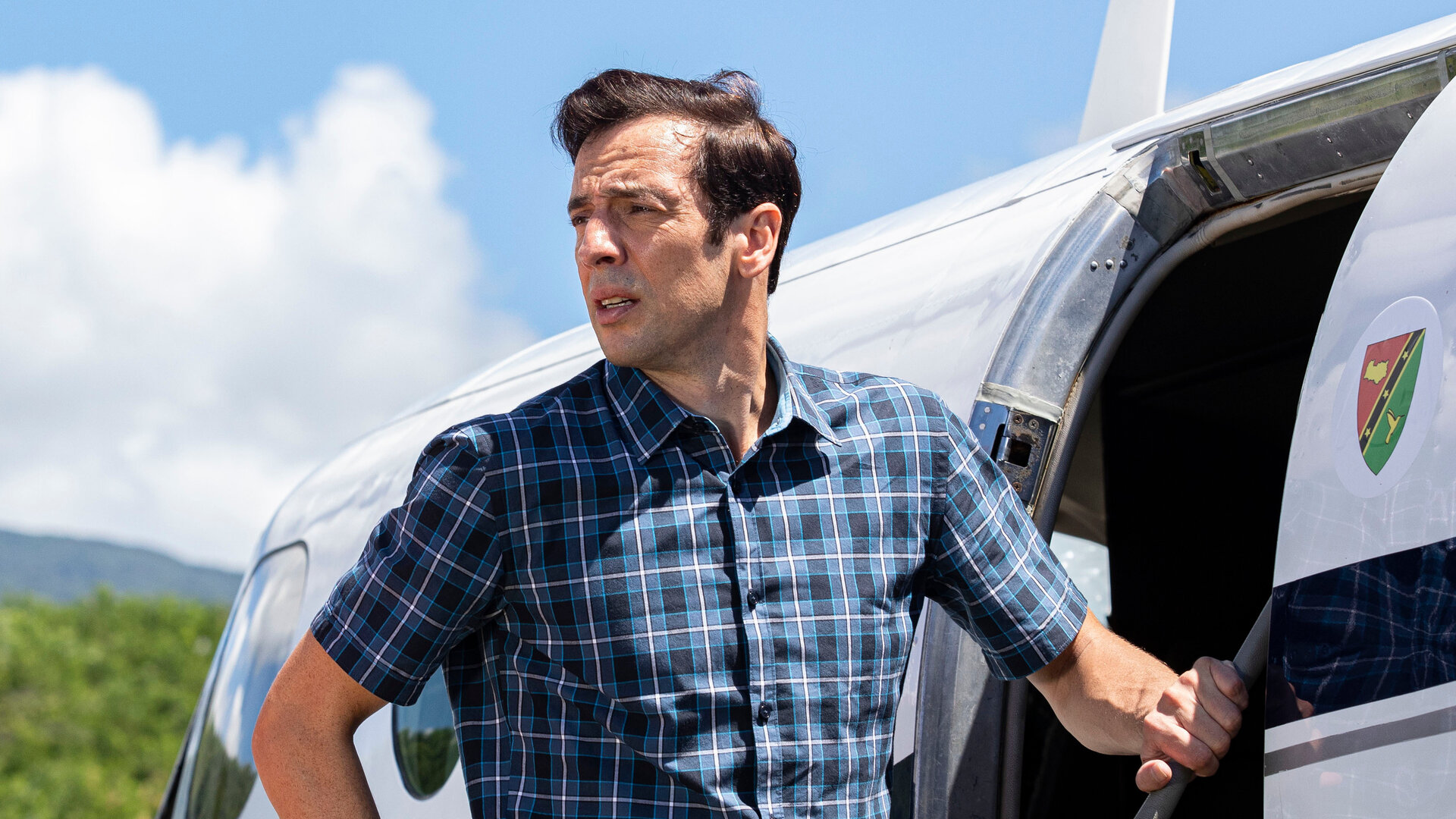 "Death in Paradise - Saint Auguste": DI Neville Parker (Ralf Little) steht nachdenklich vor einer Flugzeugtür und blickt seitlich in die Ferne.