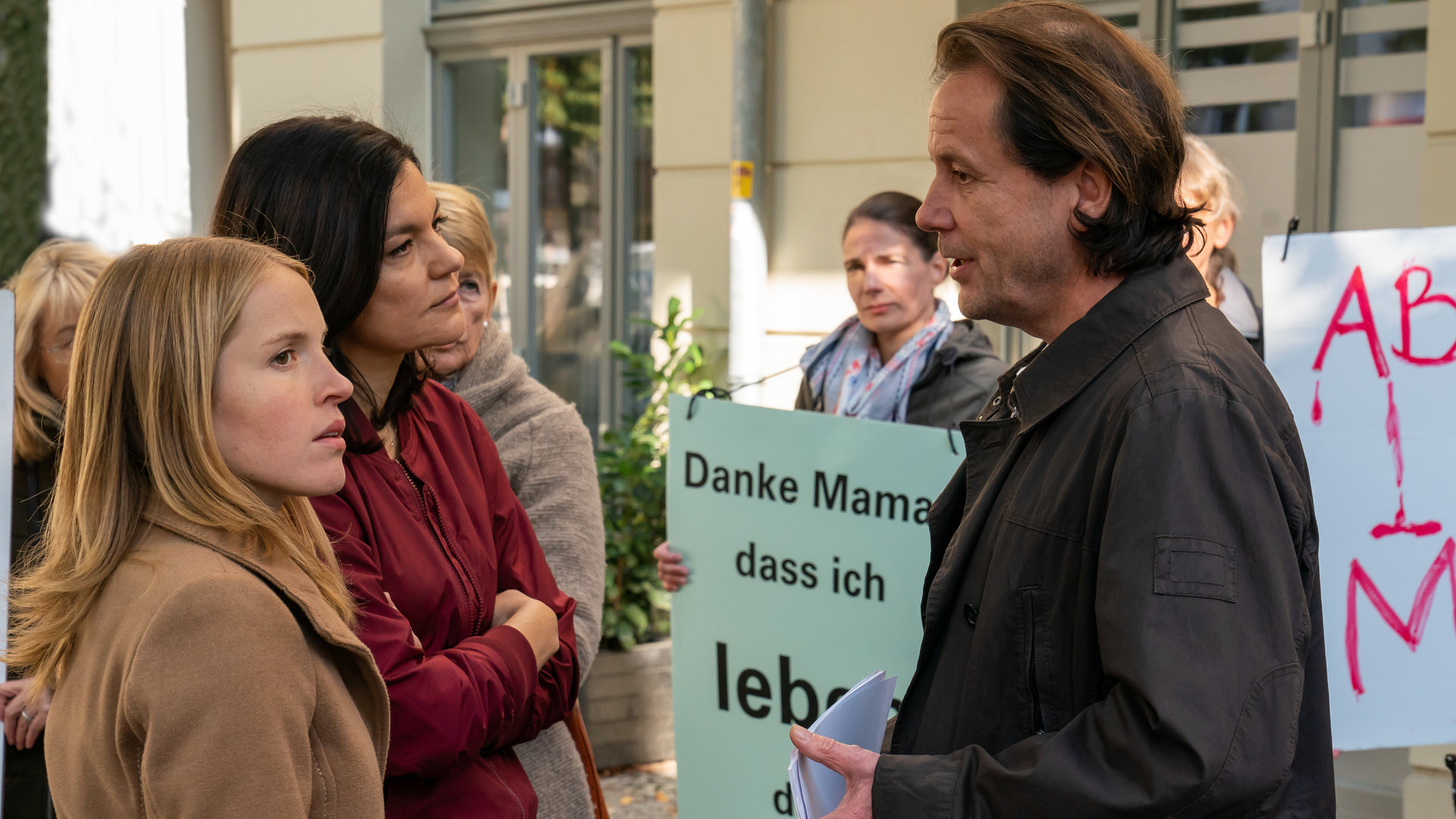 "Letzte Spur Berlin - Taufpatin": Jessica Papst (Paula Kalenberg) und Mina Amiri (Jasmin Tabatabai) stehen nebeneinander vor Martin Lipp (Christian Kuchenbuch). Um sie herum stehen mehrere Frauen mit Plakaten.