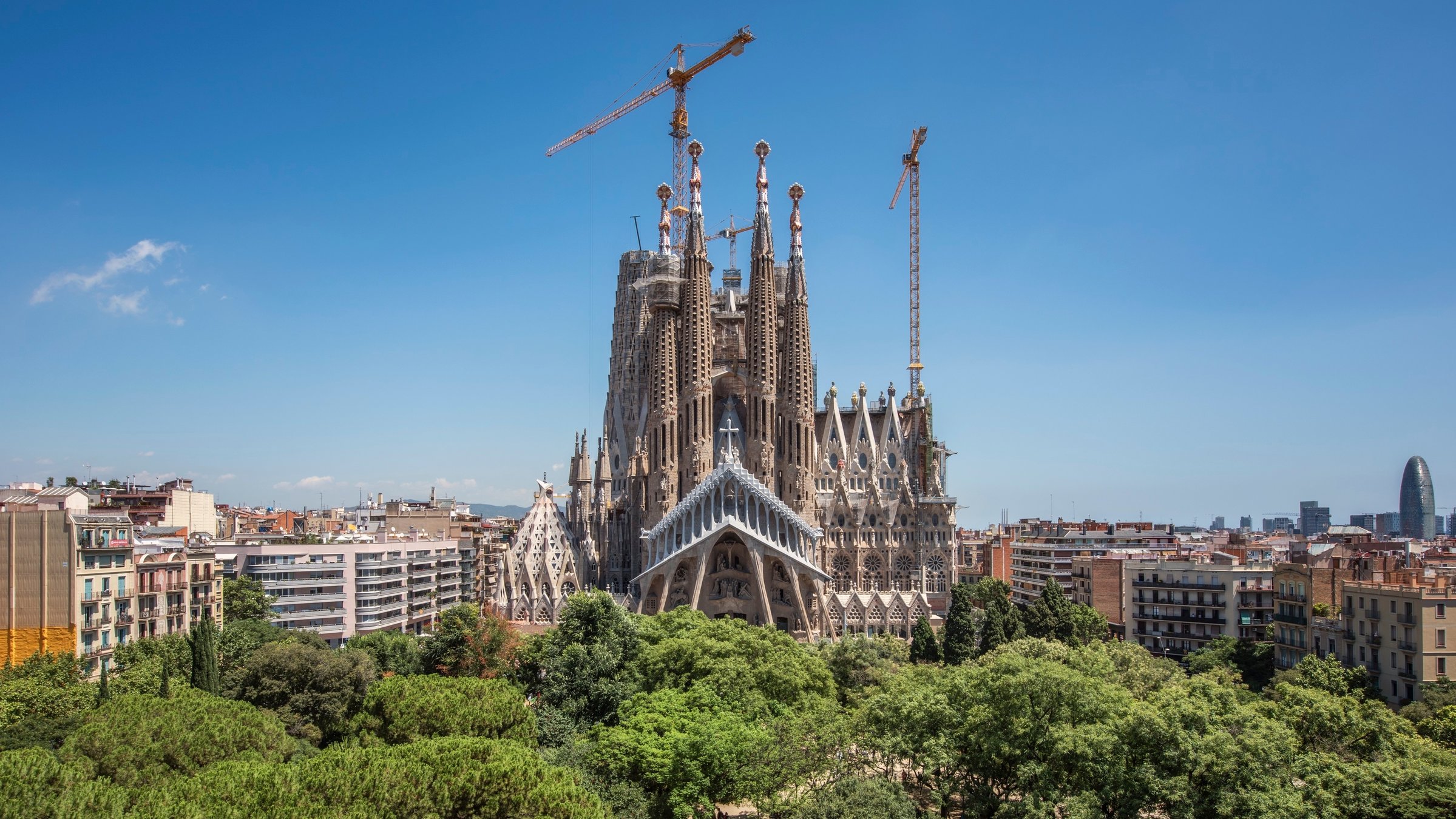 "Konzert der Wiener Philharmoniker": Außenansicht der Sagrada Familia. Die berühmte Basilika des spanischen Architekten Antoni Gaudí befindet sich bereits seit 1882 im Bau. Besonders auffallend sind die zahlreichen Türme. Zu sehen außerdem einige Baukräne und im Vordergrund viele Bäume.