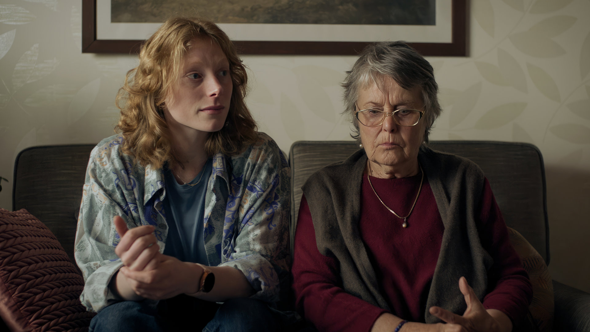 "SOKO Stuttgart - Enkeltrick": Fiona Kimmerle (Dana Herfurth) und Irmgard Löffler (Petra Kelling) sitzen auf einem alten grauen Sofa. Fiona blickt sorgenvoll zur Seite, Irmgard hat den Blick gedankenverloren gesenkt.
