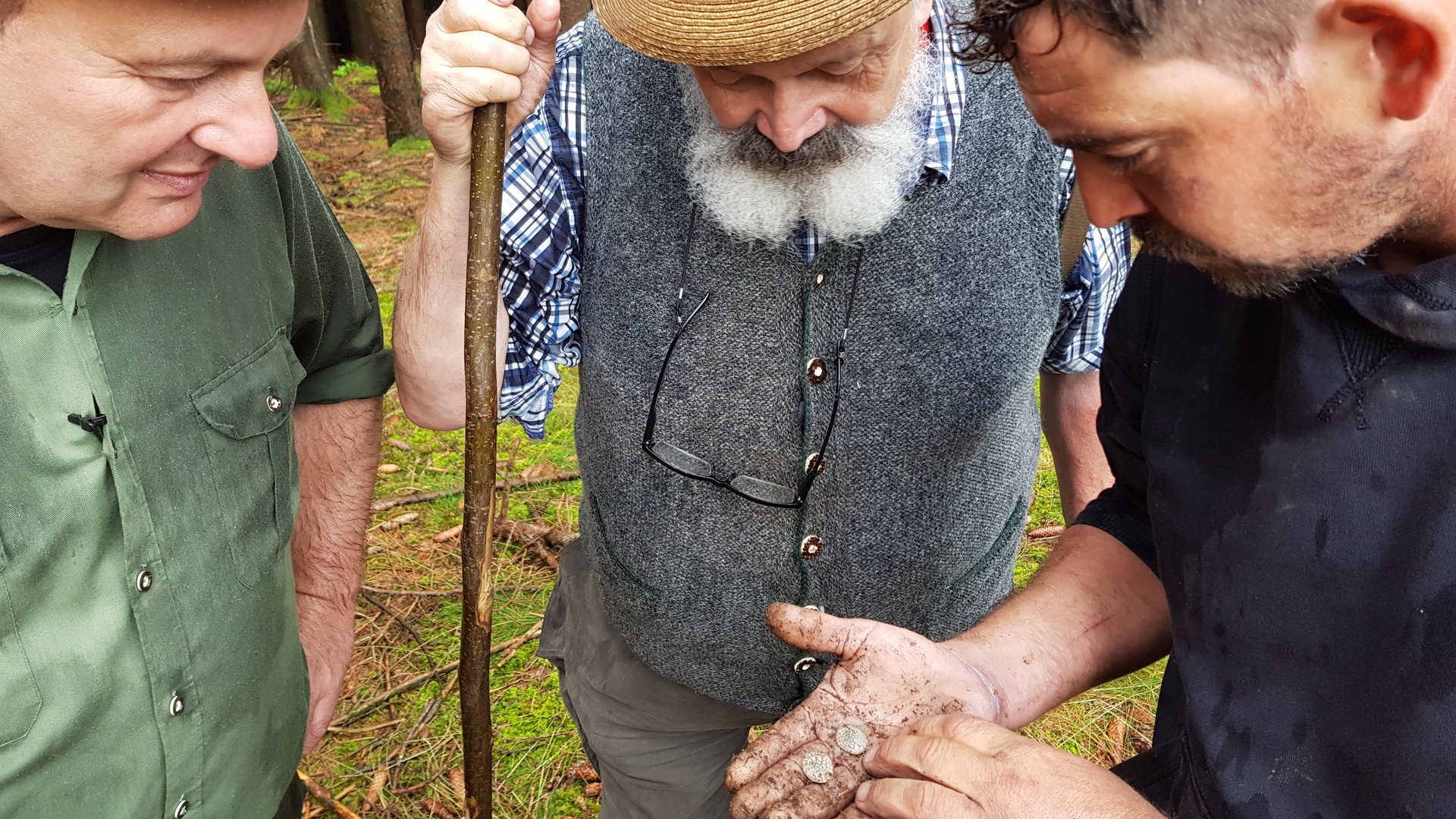 "Terra Xpress: Geraubte Schätze und eine falsche Liebe": Drei Männer stehen im Wald eng zusammen. Einer von ihnen hat ein paar Münzen in seiner offenen, verschmutzen Hand. Alle Männer schauen darauf.