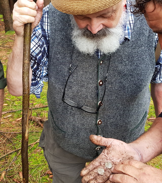 "Terra Xpress: Geraubte Schätze und eine falsche Liebe": Drei Männer stehen im Wald eng zusammen. Einer von ihnen hat ein paar Münzen in seiner offenen, verschmutzen Hand. Alle Männer schauen darauf.