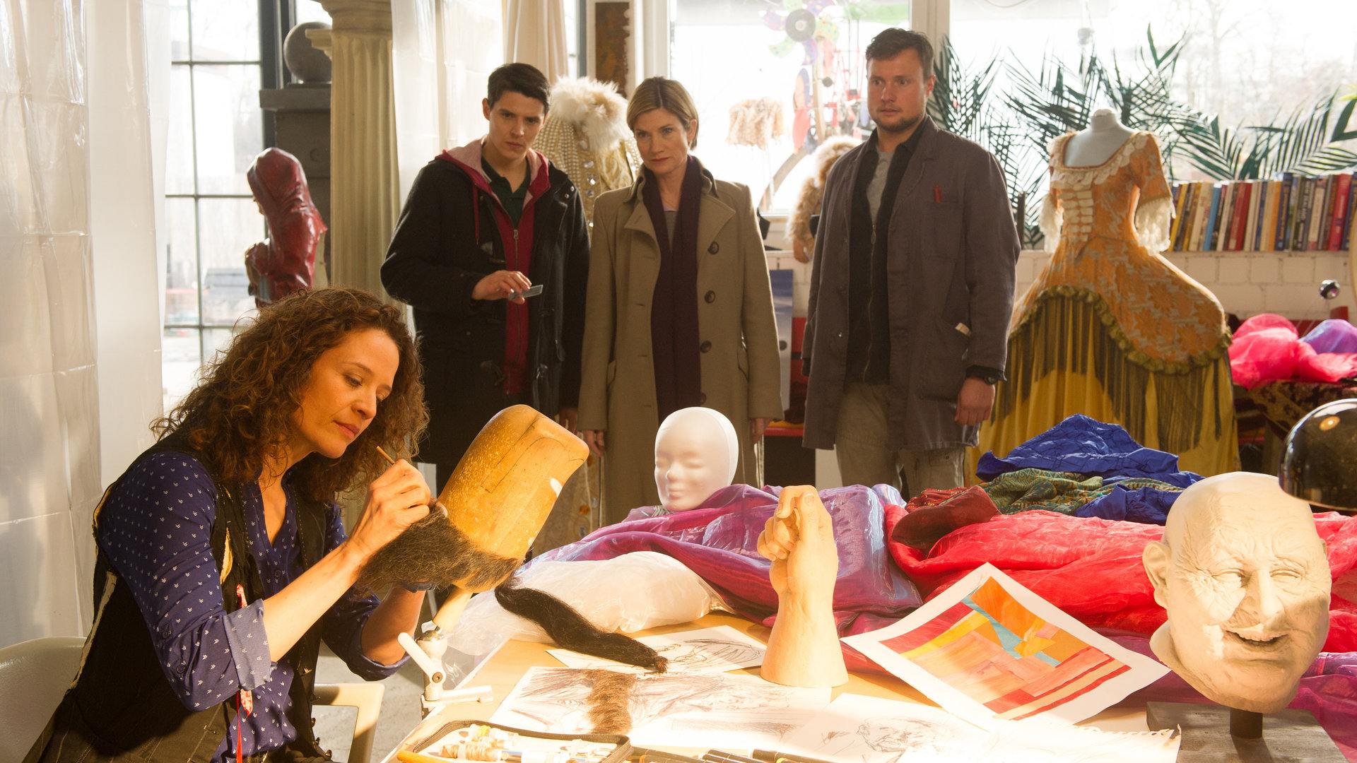 "SOKO Stuttgart - Masken": Martina Seiffert (Astrid M. Fünderich), Rico Sander (Benjamin Strecker) und Kai Wagner (Maximilian Laprell) stehen im Atelier der Bühnenbildnerin Maren Sommer (Regula Grauwiller). Diese sitzt am Tisch und klebt Haare an eine Maske. Bunte Stoffe und Masken liegen und stehen herum.