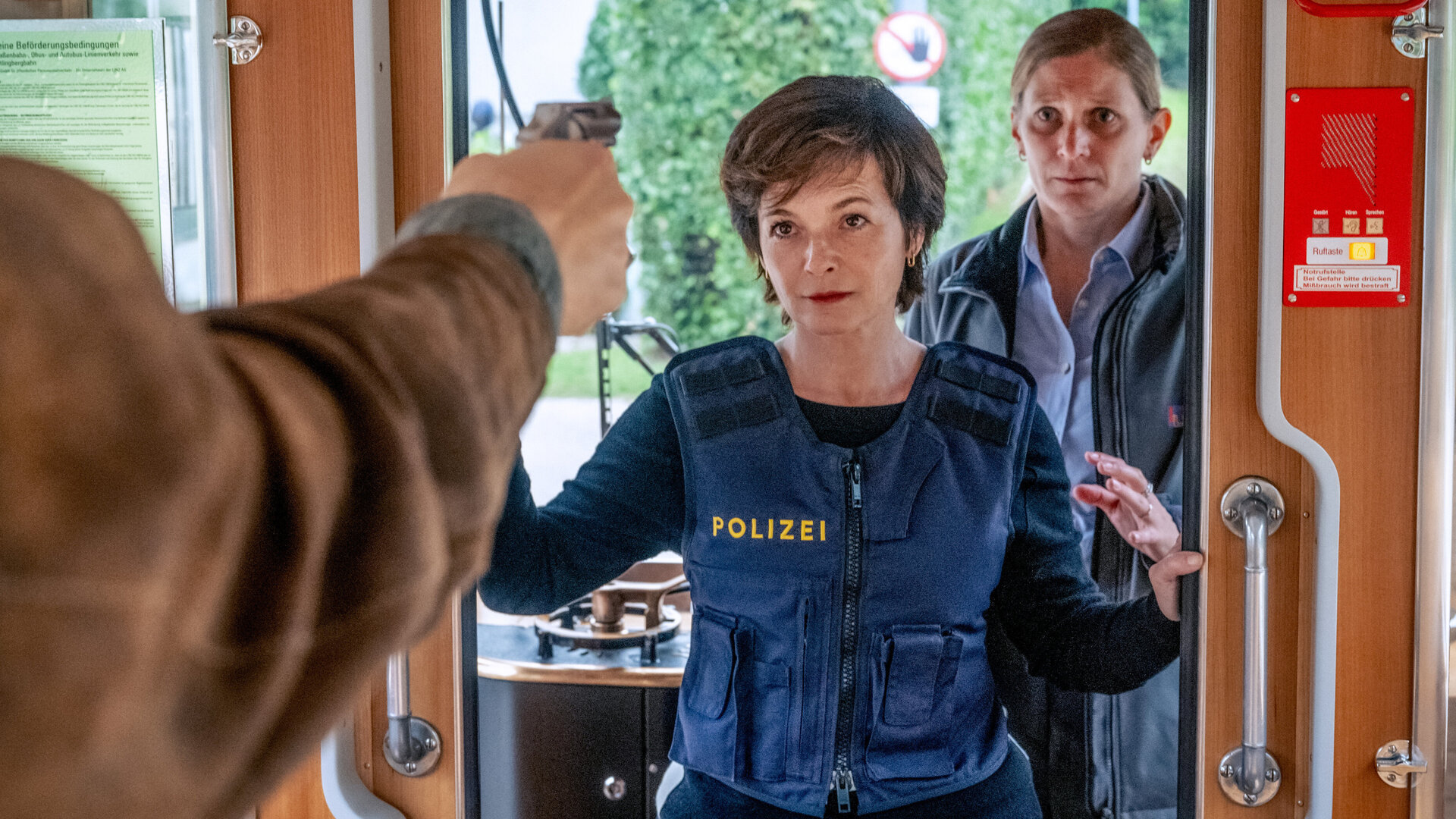 "SOKO Linz - Weichenstellung": Sven Paulitsch (Matthias Lier), der nur im Anschnitt zu sehen ist, hält eine Pistole auf Joe (Katharina Stemberger) gerichtet, die eine Schutzweste trägt und sich im Führerhaus einer Straßenbahn befindet. Birgit Sambauer (Magdalena Kronschläger) steht hinter Joe und blickt ängstlich auf die Waffe.
