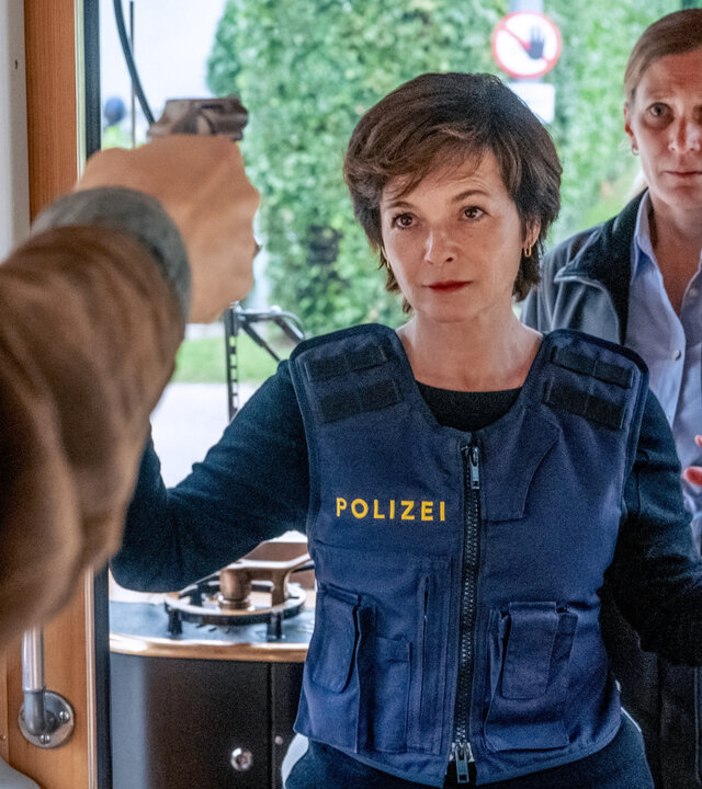 "SOKO Linz - Weichenstellung": Sven Paulitsch (Matthias Lier), der nur im Anschnitt zu sehen ist, hält eine Pistole auf Joe (Katharina Stemberger) gerichtet, die eine Schutzweste trägt und sich im Führerhaus einer Straßenbahn befindet. Birgit Sambauer (Magdalena Kronschläger) steht hinter Joe und blickt ängstlich auf die Waffe.