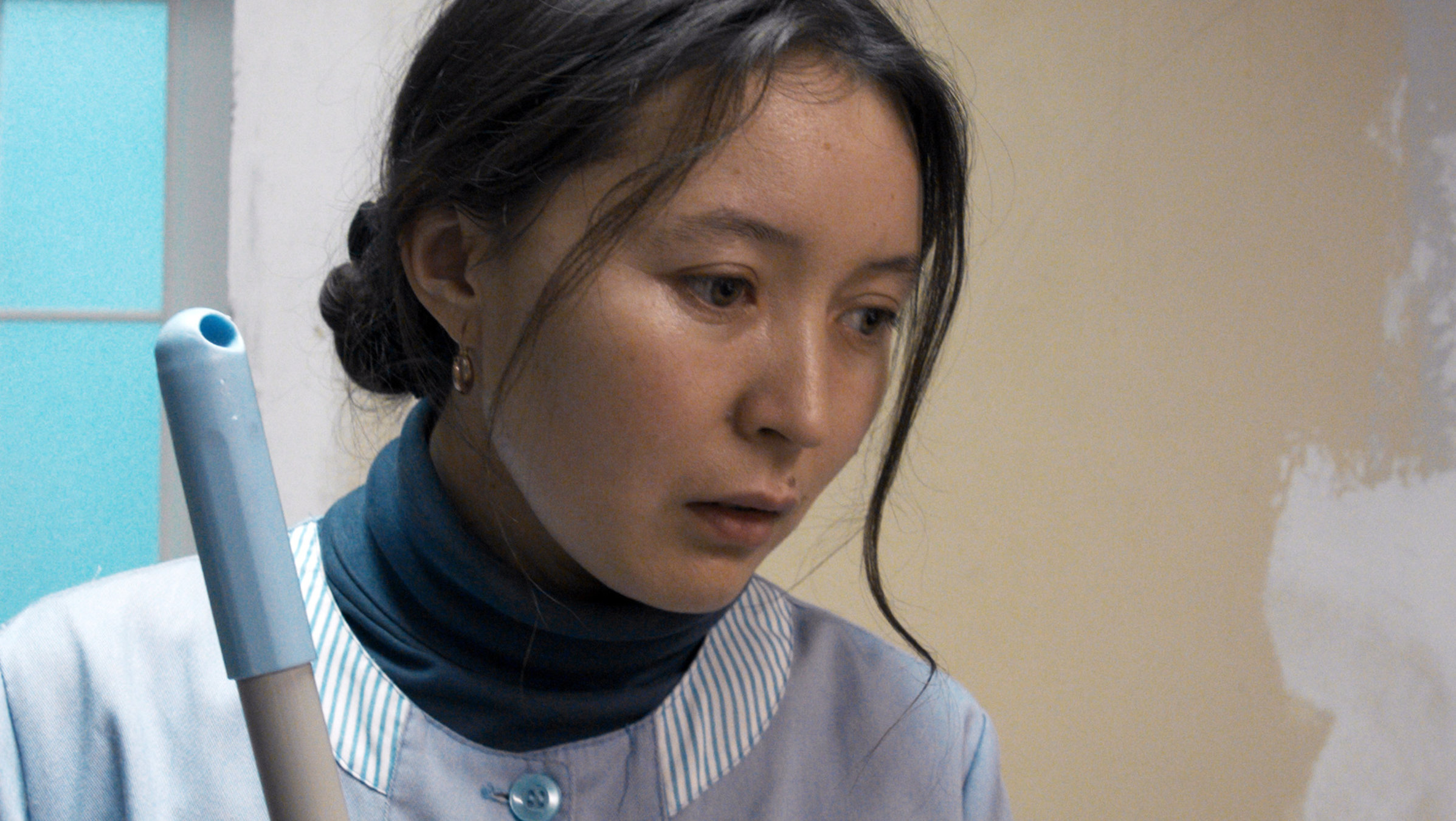 "AYKA": Ayka (Samal Yeslyamova) trägt einen grauen Putzkittel und schaut traurig zu Boden.