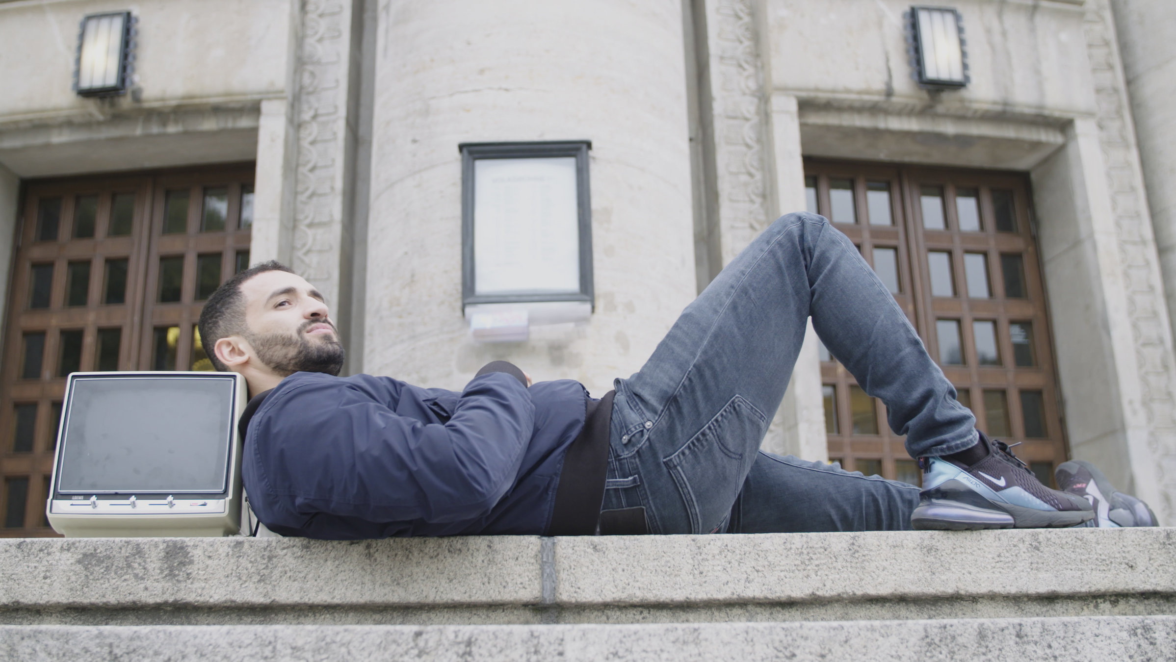 "Kino Kanak. Warum der deutsche Film Migranten braucht": Der Schauspieler Hassan Akkouch liegt vor einem Gebäude auf einer Treppe. Seinen Kopf hat er auf einem alten Fernseher abgelegt und blickt seitlich in die Ferne.