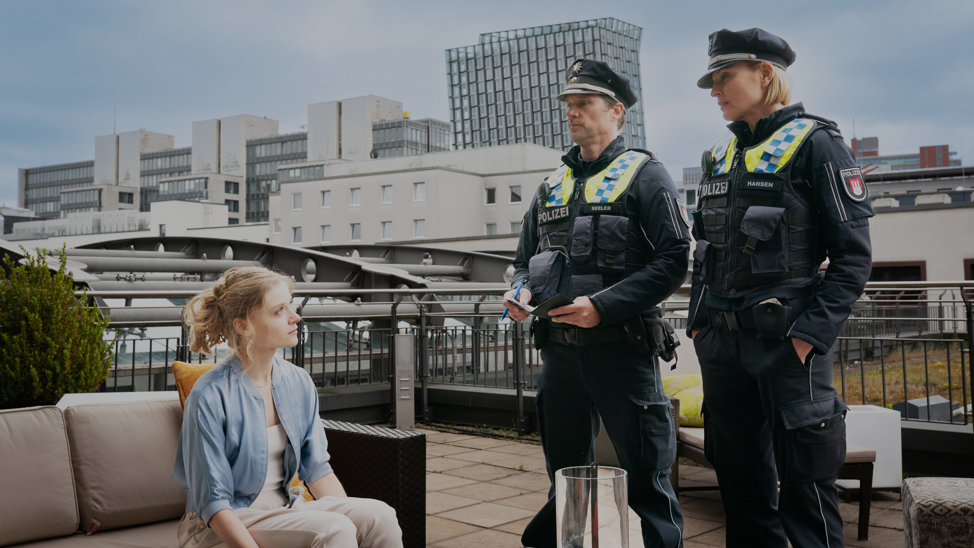 "Notruf Hafenkante - Im Netz der Lügen": Der Polizist Mattes Seeler (Matthias Schloo) und seine Kollegin Melanie Hansen (Sanna Englund) stehen nebeneinander auf der Terrasse eines Penthouses. Vor ihnen auf einem Sofa sitzt Sarah (Lara Feith). Sie blickt die beiden an. Im Hintergrund liegt eine Skyline mit weiteren Hochhäusern.
