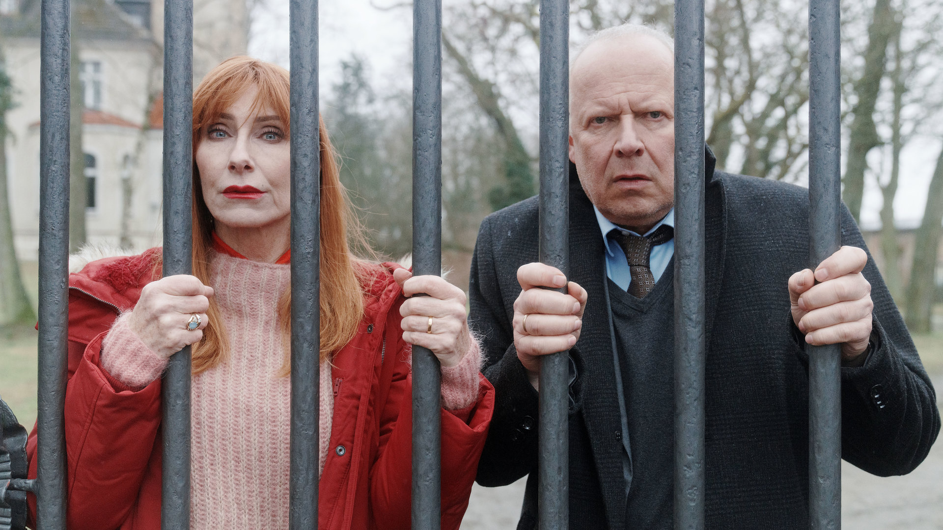 "Familie Bundschuh - Unter Verschluss": Gerald ( Axel Milberg) und Gundula (Andrea Sawatzki) krallen sich an ihrem Eisenzaun fest und blicken hilflos durch die dicken Gitterstäbe. Im Hintergrund ist der großangelegte Garten und ein Teil des Gutshauses zu sehen.