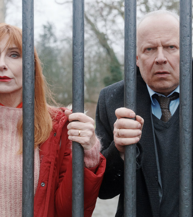 "Familie Bundschuh - Unter Verschluss": Gerald ( Axel Milberg) und Gundula (Andrea Sawatzki) krallen sich an ihrem Eisenzaun fest und blicken hilflos durch die dicken Gitterstäbe. Im Hintergrund ist der großangelegte Garten und ein Teil des Gutshauses zu sehen.